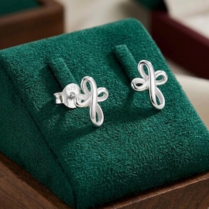 May include: A pair of silver stud earrings featuring a stylised cross design with looped, continuous lines. The back of one earring is clearly stamped with the number 925, indicating the silver content. The earrings have a polished, metallic finish.