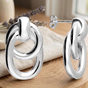 May include: A pair of silver earrings with a linked oval design. The earrings have a post for pierced ears. They are displayed on a wooden surface with a neutral-coloured cloth. The earrings are made of a shiny metal.