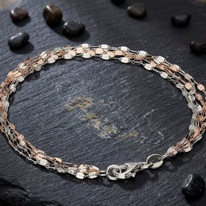 Könnte beinhalten: Ein mehrsträngiges Armband mit einer Mischung aus Silber- und Roségoldtönen. Das Armband hat ein filigranes, gewebtes Design mit einem sicheren Verschluss. Der Schmuck wird auf einer dunklen, strukturierten Oberfläche mit kleinen, dunklen Steinen präsentiert.