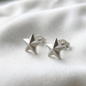 Peut inclure: Une paire de boucles d'oreilles à clous en forme d'étoile en argent avec une finition en métal brossé. Chaque boucle d'oreille présente un design à cinq branches avec une arête centrale et un fermoir en argent marqué du nombre 925.