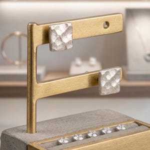 Könnte beinhalten: Ein Schmuckständer mit zwei silbernen, quadratischen Ohrringen mit strukturierter Oberfläche. Die Ohrringe sind an einem goldfarbenen Balken befestigt. Der Ständer hat auch eine graue Basis mit einer Reihe kleiner, klarer Edelsteine.