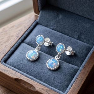 May include: A pair of silver drop earrings featuring two round, iridescent blue opal cabochons per earring. Each stone is set in a beaded silver frame, with the top stone serving as the stud and the bottom stone dangling below.