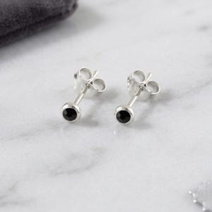 May include: A pair of silver stud earrings, each featuring a small, round, black gem. The earrings are set against a white marble surface, with a grey pouch visible.