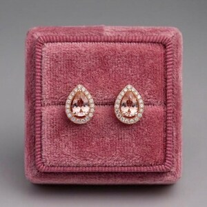 May include: A pair of teardrop-shaped earrings featuring a central peach-colored gemstone surrounded by a halo of small white stones, set in a rose-colored metal frame. The earrings are displayed on a square, deep pink velvet jewelry box.