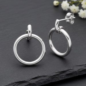 May include: A pair of polished silver hoop stud earrings. Each earring has a circular hoop design attached to a post with a butterfly clasp. The earrings are presented on a dark, textured surface.