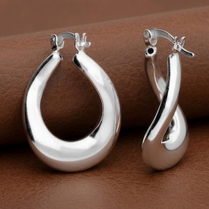 May include: Two silver hoop earrings. One is a simple teardrop shape, the other has a twisted design. The earrings are highly polished, reflecting light and appearing bright. The background is a solid brown colour. The earrings are likely made of metal.