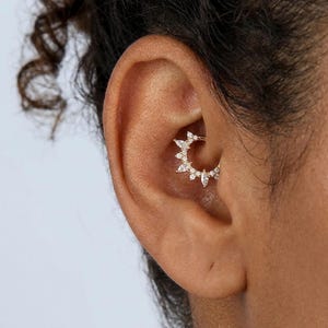 Può includere: Un anello daith in oro con una disposizione semicircolare di pietre trasparenti a taglio marquise e rotondo. Il gioiello è posizionato all'interno della cartilagine dell'orecchio, mostrando un design a raggiera con molteplici gemme incastonate.
