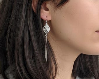 Könnte beinhalten: Silberne Filigran-Ohrringe mit Tropfendesign und hängenden Ketten. Die Ohrringe werden von einer Person mit dunklem Haar und einem grauen Pullover getragen. Das filigrane Muster und die zarten Ketten erzeugen einen eleganten Look.