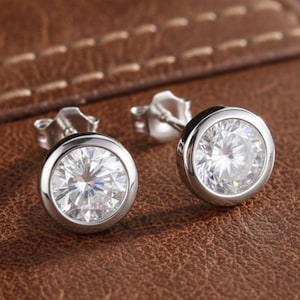 May include: Two silver stud earrings, each featuring a large, clear, round gemstone. The gemstones are set in a bezel setting. The earrings are presented on a brown leather surface.