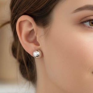 May include: A close-up of an ear with a silver ball stud earring. The earring is a simple, polished sphere, reflecting light. The skin tone is fair, and the hair is brown.