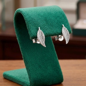 May include: A pair of silver stud earrings shaped like delicate leaves with detailed vein textures. Each earring features a polished metallic finish and a secure post backing, showcasing a slender, pointed design that mimics natural foliage.