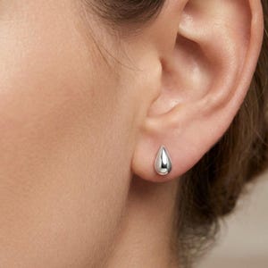 May include: A small, polished silver stud earring shaped like a smooth, rounded teardrop. The earring features a reflective, metallic surface and is secured directly against the earlobe, presenting a simple, minimalist, and clean geometric form.