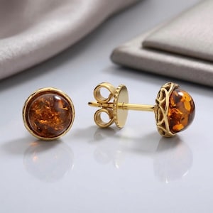 May include: A pair of stud earrings featuring round, polished amber cabochons set in gold-toned metal. The settings have a decorative, openwork filigree pattern around the sides, and the earrings include standard post backs with secure metal clutches.