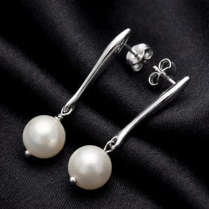 Può includere: Un paio di orecchini pendenti in argento con una perla bianca all'estremità. Gli orecchini hanno un design elegante e allungato, su sfondo nero.