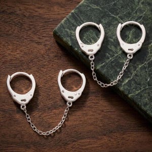 May include: A pair of silver-toned hoop earrings, each featuring two circular hoops connected by a short, delicate silver chain. The design mimics the appearance of miniature handcuffs, with a polished metallic finish and a hinged clasp mechanism.