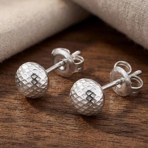 May include: A pair of silver stud earrings. Each earring features a textured, spherical design. The earrings are displayed on a wooden surface with a neutral-colored fabric in the background. The earrings are small and delicate.