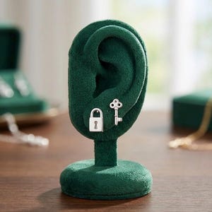 May include: A pair of silver-toned stud earrings featuring a padlock design and a key design. The earrings are displayed on a dark green, velvet-textured ear-shaped stand. The padlock earring is rectangular with a keyhole, and the key earring is shaped like a classic skeleton key with a round bow.