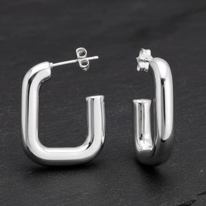 Könnte beinhalten: Ein Paar glänzende, silberfarbene quadratische Creolen. Die Ohrringe haben einen Stecker und einen Butterfly-Verschluss. Die Ohrringe werden auf einer dunklen Oberfläche präsentiert. Modernes Schmuckdesign.