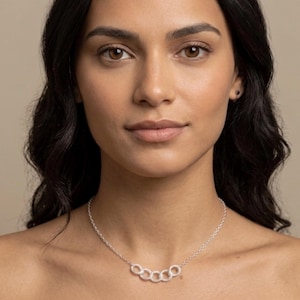 Puede incluir: Un collar plateado con cinco círculos interconectados. El collar lo lleva una persona con cabello oscuro y ojos marrones. El fondo es de un color neutro sólido.