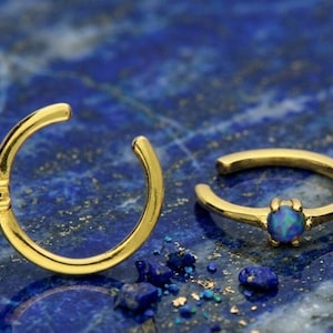 May include: Two gold ear cuffs, each featuring a small, oval, blue gemstone. The cuffs are open at one end. The background is a dark blue, textured surface with small blue and gold flecks.
