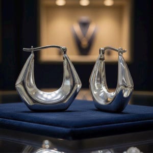 May include: A pair of silver hoop earrings with a geometric, faceted design. The earrings have a polished, reflective surface and a hinged closure. They are displayed on a dark blue velvet surface, with a blurred background of jewelry.