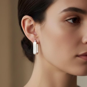 May include: Silver rectangular hoop earrings. The earrings have a simple, modern design with a polished finish. The earrings are shown on a person's ear, with a soft background.