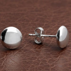May include: A pair of shiny, silver-colored ball stud earrings. One earring is a complete sphere, while the other is an oval shape. The post of the earring is visible, with a small clasp. The earrings are set against a brown leather-textured background.