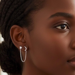 May include: Silver chain earrings featuring a star and crescent moon design. The star is on the top and the moon is on the bottom, connected by a delicate chain. The earrings are worn on the ear.