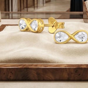 Puede incluir: Un par de pendientes de botón dorados con un diseño de símbolo de infinito. Cada pendiente está engastado con dos piedras transparentes en forma de pera dentro del marco dorado. Los pendientes incluyen cierres de presión dorados.