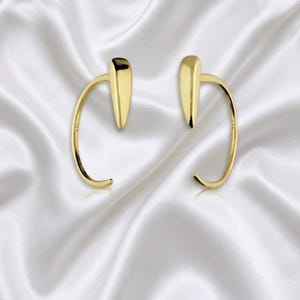 May include: A pair of gold-toned ear cuffs featuring a smooth, curved wire design that wraps around the earlobe, ending in a pointed, teardrop-shaped accent at the top. The polished metal surface reflects light, creating a bright, metallic appearance.
