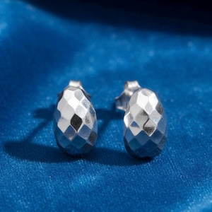 May include: A pair of silver-colored stud earrings with a faceted, teardrop design. The earrings are displayed on a blue velvet surface, reflecting light and creating a sparkling effect. The earrings have a post backing.
