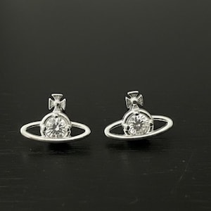 May include: A pair of silver-toned stud earrings shaped like the planet Saturn with a ring around the center. Each earring features a clear, round-cut gemstone at the core and a small cross emblem extending from the top of the planetary sphere.