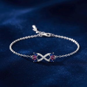 Puede incluir: Una pulsera de plata con un diseño de símbolo de infinito. El símbolo del infinito está adornado con pequeñas piedras preciosas claras, azules y rosas. La pulsera está sobre un fondo azul oscuro.