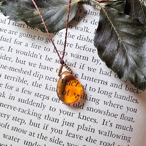 May include: A teardrop-shaped amber-colored pendant suspended from a thin brown cord. The pendant features a small cluster of metallic gold-toned beads at the top where it connects to the cord, creating a simple and delicate jewelry piece.