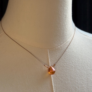May include: A delicate necklace featuring a single teardrop-shaped orange gemstone pendant suspended on a thin metallic chain. The pendant is flanked by two small, matching orange beads on either side, creating a simple and refined jewelry piece.