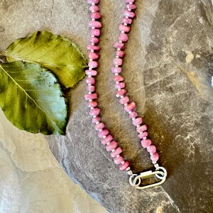 May include: A pink gemstone necklace with a silver clasp. The necklace is made of small, irregularly shaped pink stones strung together. The clasp is a silver oval shape. A green leaf is in the background.