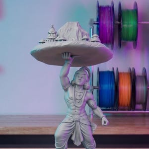 May include: A grey figurine of a muscular man supporting a large, flat, circular object above his head. The object features a mountain and structures. The man wears traditional clothing and jewellery. Spools of colourful filament are in the background.