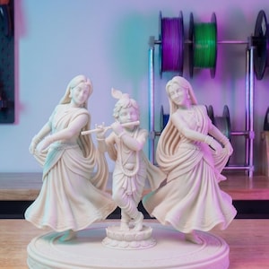 Può includere: Una scultura color crema di tre figure: una figura centrale che suona il flauto, affiancata da due figure danzanti. Le figure sono su una base circolare. Lo sfondo include bobine di filamento.