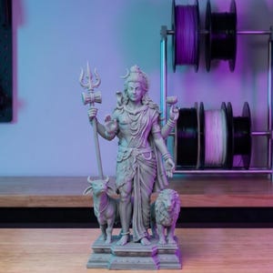 Puede incluir: Una estatua gris de una deidad sosteniendo un tridente y una flor de loto, flanqueada por un toro y un león. La estatua está sobre una base rectangular. En el fondo hay varias bobinas de filamento para impresión 3D.