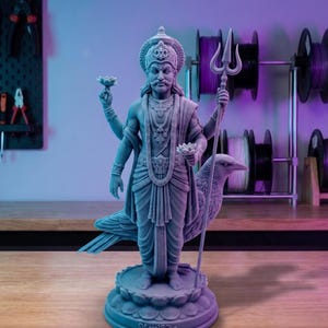 May include: A gray statue of a deity holding a trident and lotus flowers, with a raven at its feet. The figure is adorned with detailed jewelry and a crown. The statue is on a lotus flower base.
