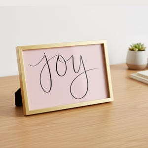 Puede incluir: Un marco dorado que contiene una impresión rosa con la palabra "joy" escrita en negro. El marco está sobre una superficie de madera. En el fondo se ve una pequeña suculenta en una maceta blanca.