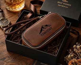 Puede incluir: Un estuche con cremallera de cuero marrón con un gráfico de montaña y el nombre "TIMOTHY" se presenta en una caja de regalo negra con las palabras "BEST WISHES" en dorado. También se ven un reloj y un vaso de whisky.