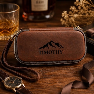 Puede incluir: Un estuche de reloj de cuero marr&oacute;n con un dise&ntilde;o de monta&ntilde;a y el nombre "TIMOTHY" impreso en negro. El estuche tiene cierre de cremallera y una correa de mu&ntilde;eca. Se ve un reloj de pulsera con una correa de cuero marr&oacute;n.