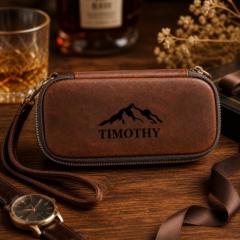 Puede incluir: Un estuche de reloj de cuero marr&oacute;n con un dise&ntilde;o de monta&ntilde;a y el nombre "TIMOTHY" impreso en negro. El estuche tiene cierre de cremallera y una correa de mu&ntilde;eca. Se ve un reloj de pulsera con una correa de cuero marr&oacute;n.