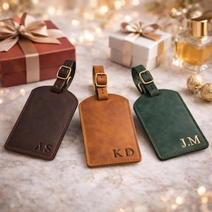 Puede incluir: Tres etiquetas de equipaje de cuero en marrón, beige y verde, cada una con una hebilla dorada e iniciales personalizadas. Las etiquetas son rectangulares con la parte superior redondeada y se muestran con regalos envueltos y luces decorativas en el fondo.