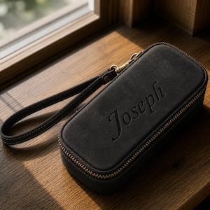 Puede incluir: Un estuche rectangular negro con cremallera dorada y correa de mu&ntilde;eca. La palabra "Joseph" est&aacute; grabada en relieve en la parte delantera. El estuche est&aacute; hecho de un material texturizado y tiene un cierre dorado.