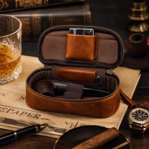 Puede incluir: Estuche de cuero marr&oacute;n abierto para pipa de tabaco con cremallera, que contiene una pipa, un encendedor y un bol&iacute;grafo. El estuche est&aacute; sobre un peri&oacute;dico, con un reloj y un vaso de l&iacute;quido &aacute;mbar en el fondo. El estuche est&aacute; dise&ntilde;ado para guardar accesorios para fumar.