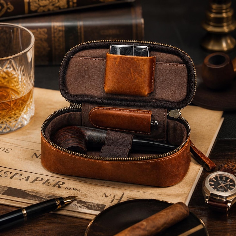 Puede incluir: Estuche de cuero marr&oacute;n abierto para pipa de tabaco con cremallera, que contiene una pipa, un encendedor y un bol&iacute;grafo. El estuche est&aacute; sobre un peri&oacute;dico, con un reloj y un vaso de l&iacute;quido &aacute;mbar en el fondo. El estuche est&aacute; dise&ntilde;ado para guardar accesorios para fumar.