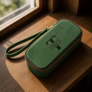 Puede incluir: Un estuche verde con cremallera y correa de mu&ntilde;eca, con el nombre "TRICE" en negro. El estuche tiene una cremallera y herrajes dorados. El estuche est&aacute; sobre una superficie de madera.