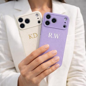 Op de afbeelding: Twee gepersonaliseerde telefoonhoesjes. Eén is gebroken wit met gouden letters "KD", de andere is lavendelkleurig met zilveren letters "R.W". Beide hoesjes hebben een textuurafwerking en een uitsparing voor de camera.